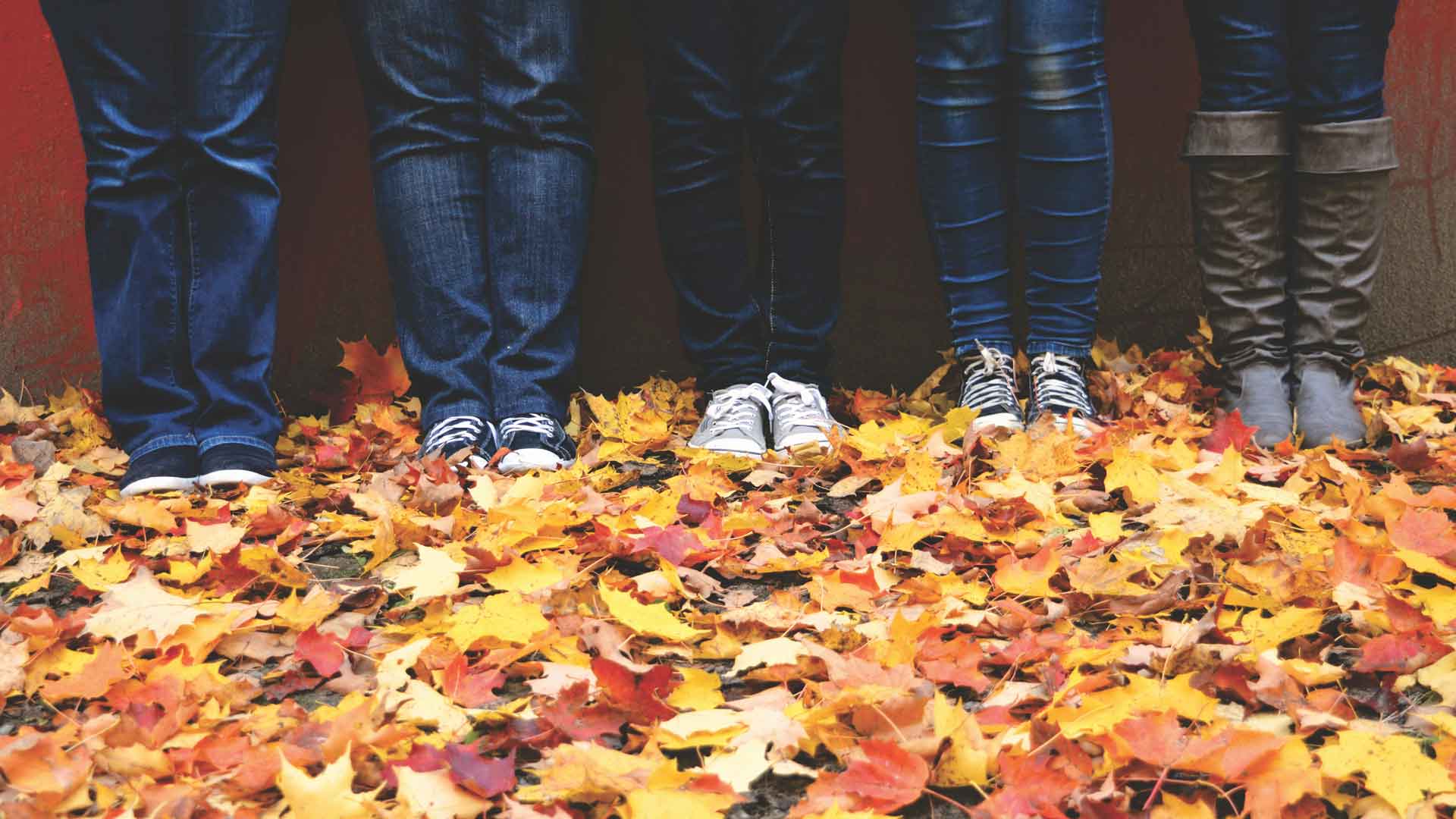 Four sets of legs with jeans and various shoes on stand next to each other in fall foliage.