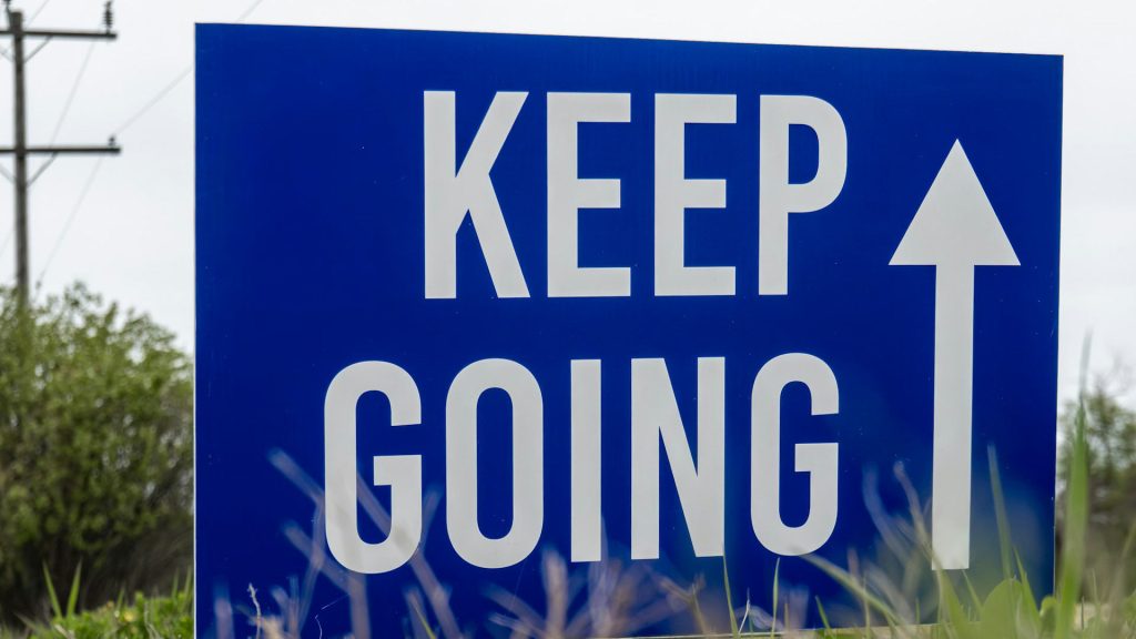 A blue sign with the words "Keep Going" printed on it in white, with a white arrow pointing straight, next to the words.